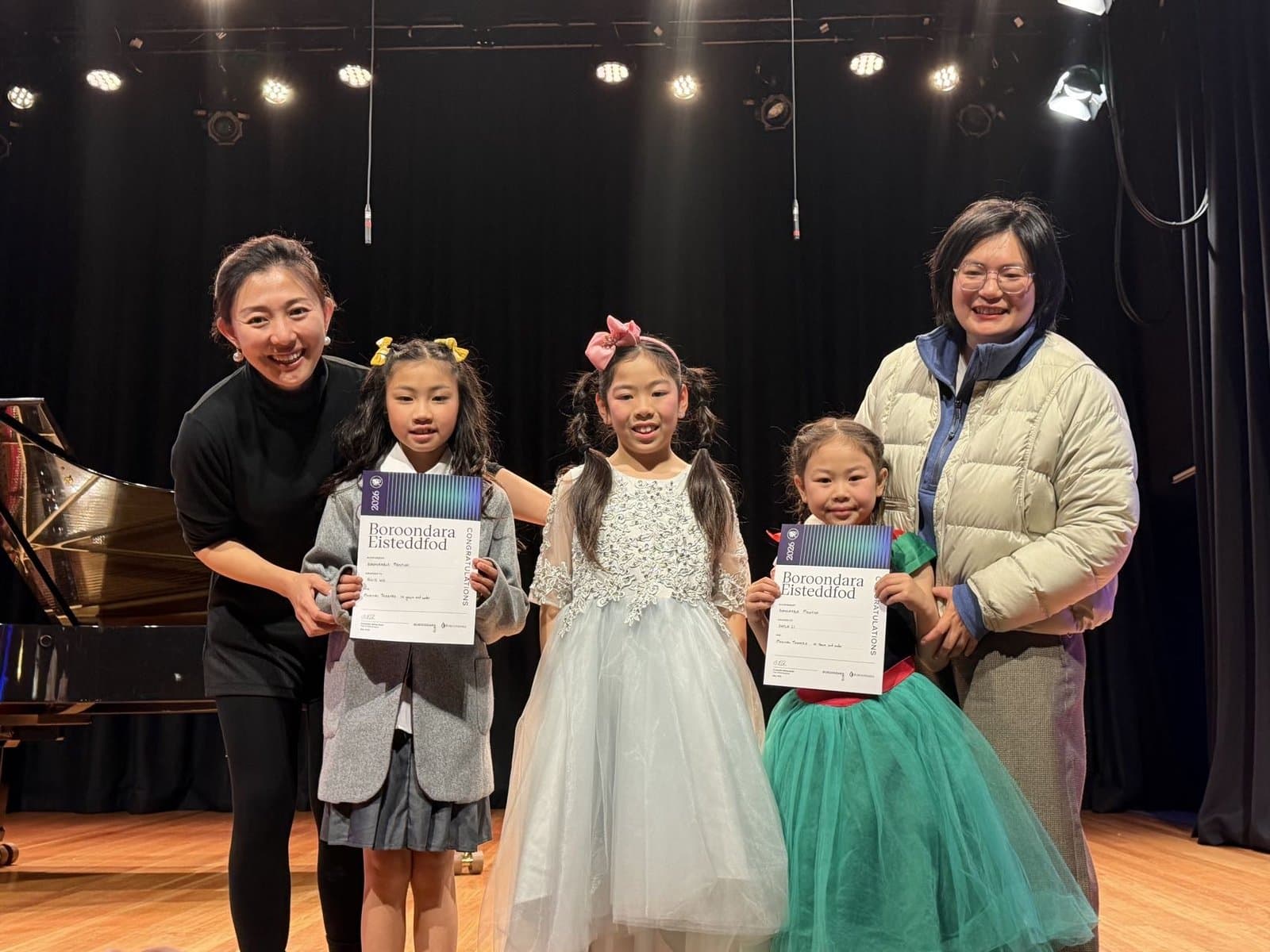 Three students holding certificates beside their teachers on a piano recital stage