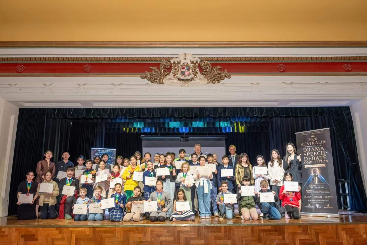 The full field of winners with certificates on the stage of a heritage hall