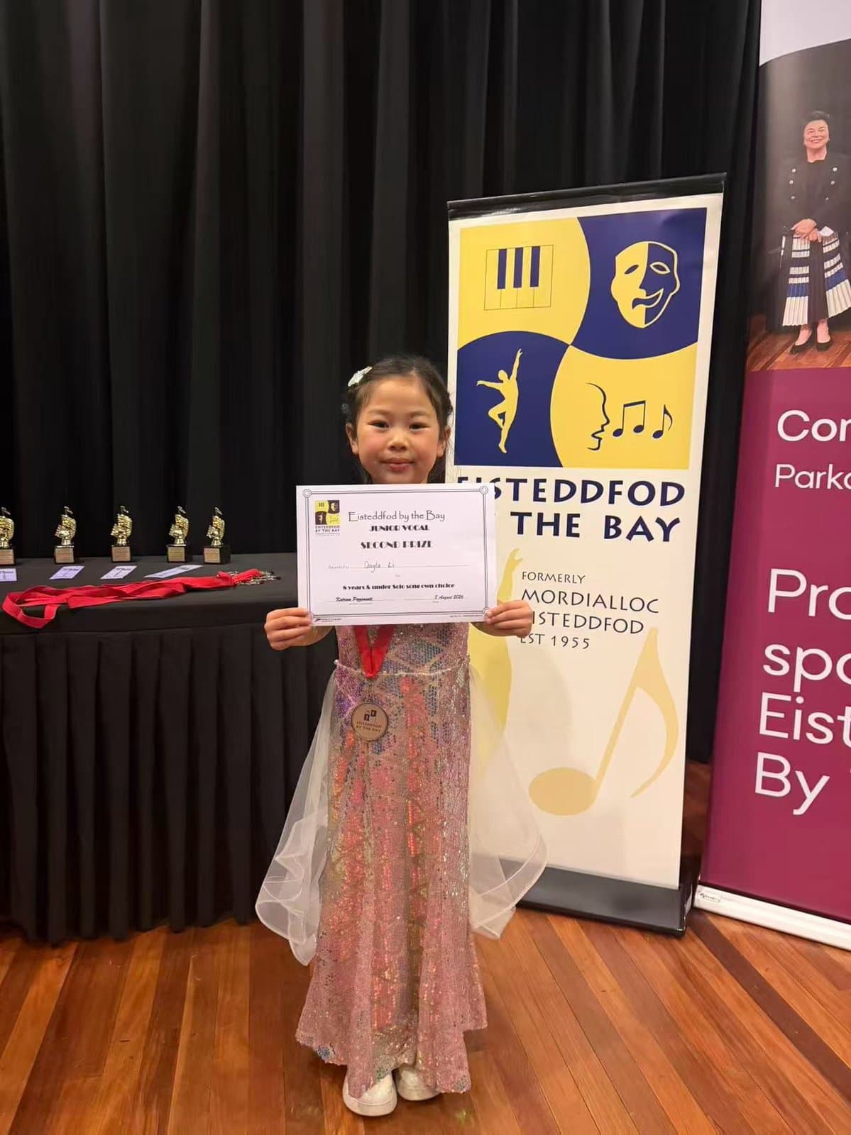 A student holding her certificate beside the Eisteddfod by the Bay banner