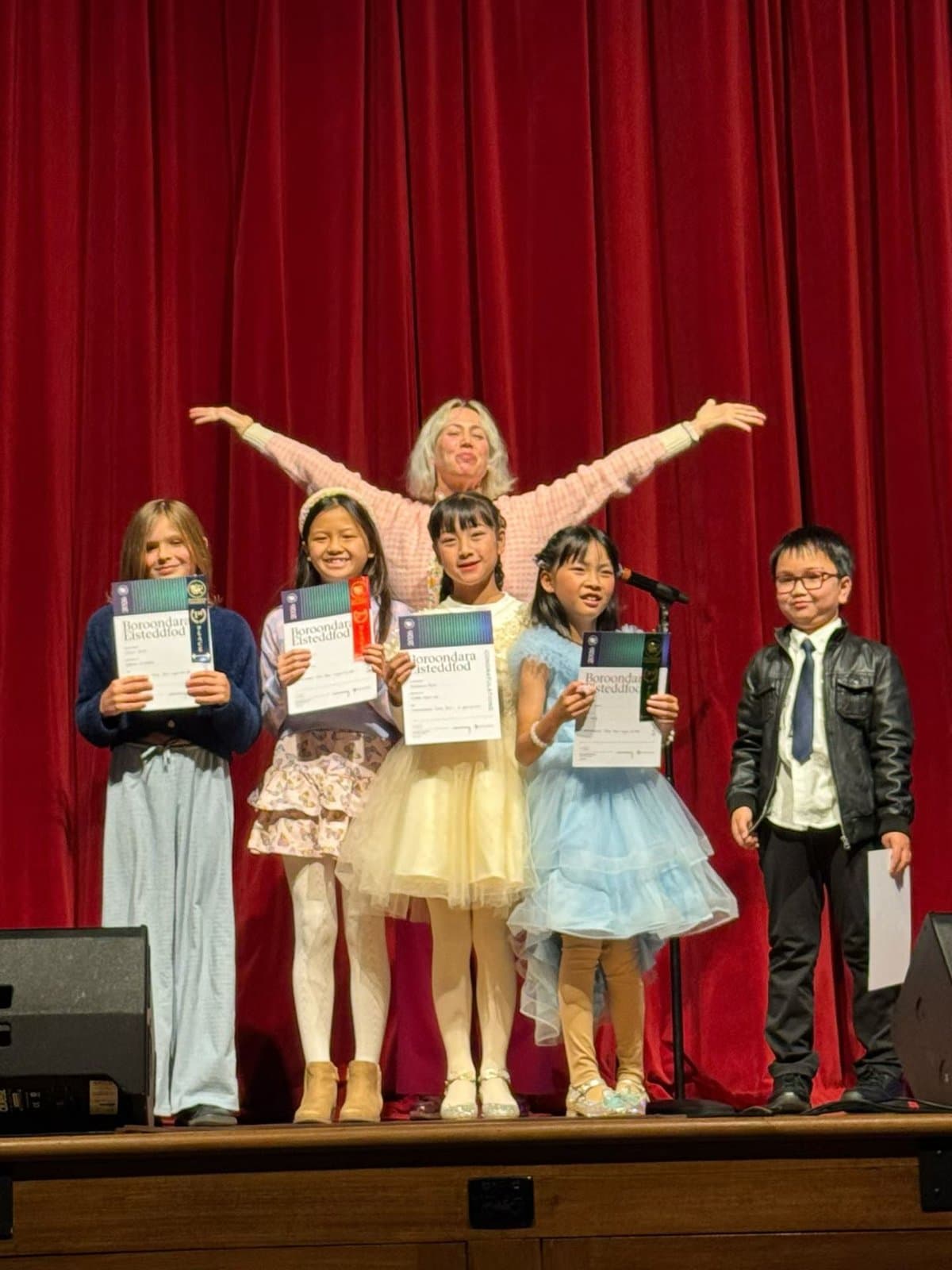 Students celebrating with certificates and their teacher on a red-curtained stage