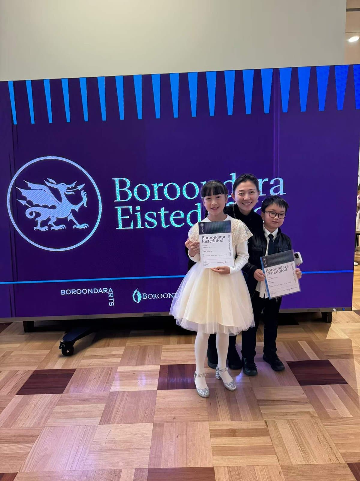 Three students holding certificates in front of the Boroondara Eisteddfod screen