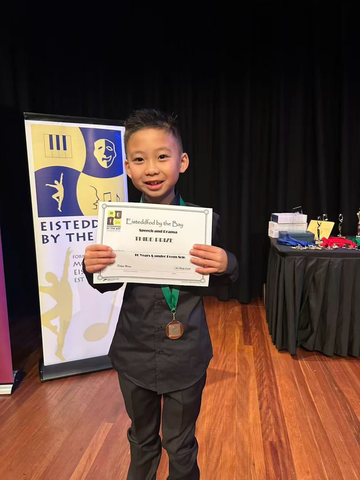 A student holding his certificate and medal beside the Eisteddfod by the Bay banner