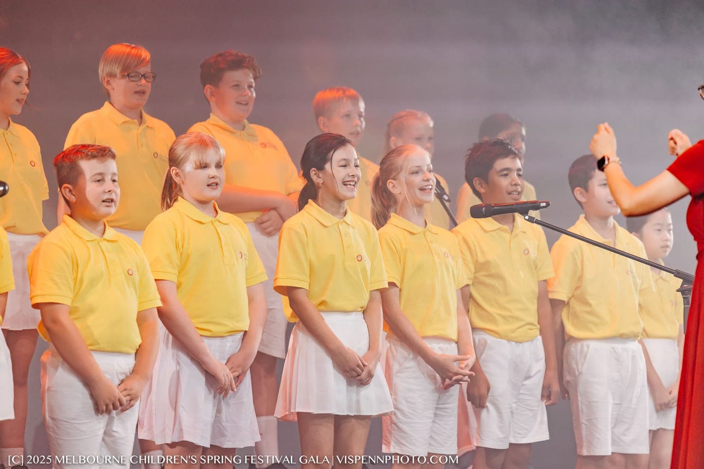 The Mirror choir in yellow polo shirts singing on the gala stage, their conductor's raised hand at the edge of the frame