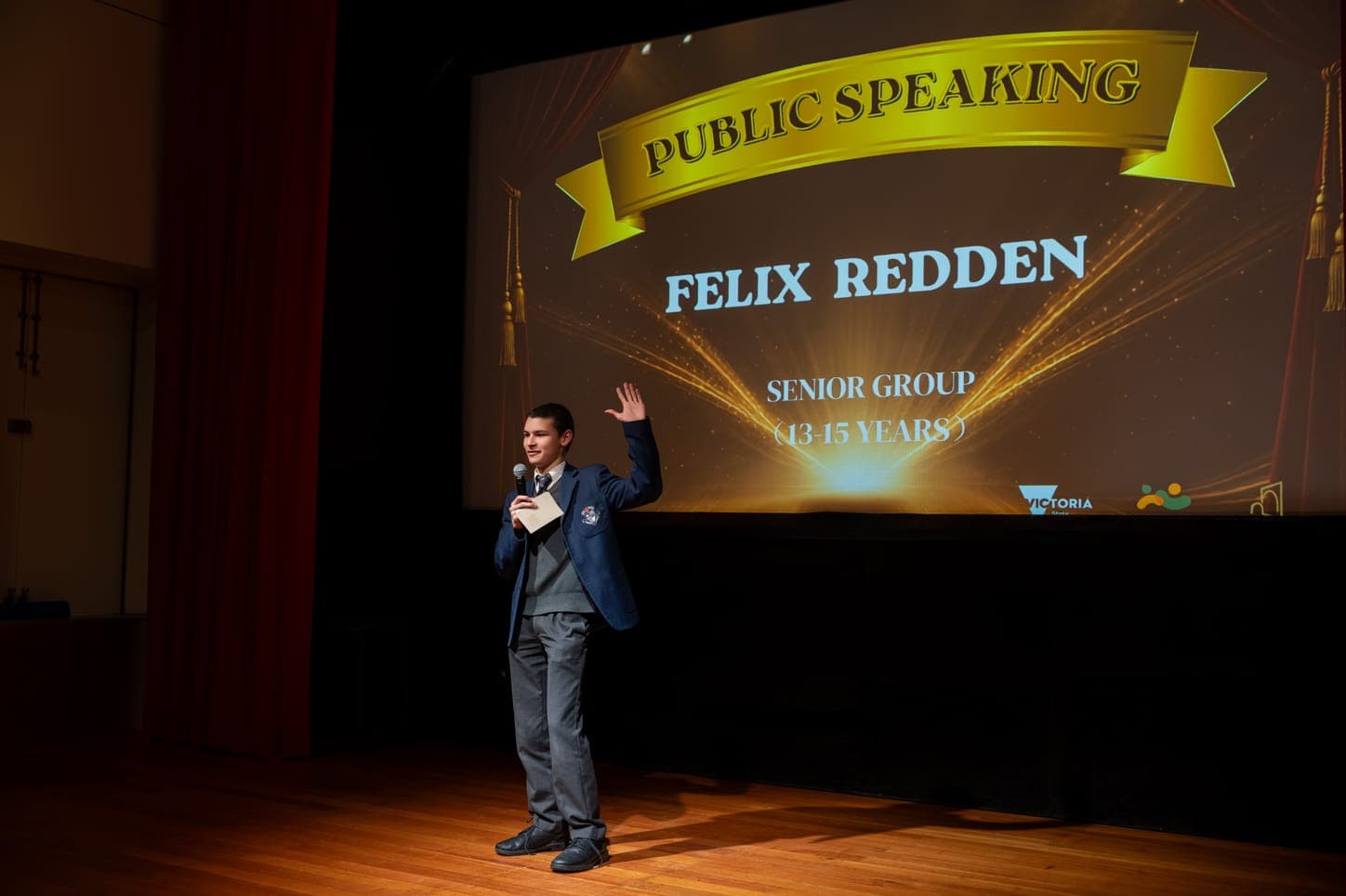 A student speaking at the microphone on a school stage, the screen behind him titled Public Speaking above the senior age group