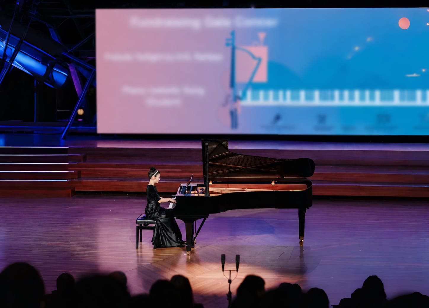 A pianist alone at a grand piano on a concert-hall stage, the audience silhouetted in the foreground
