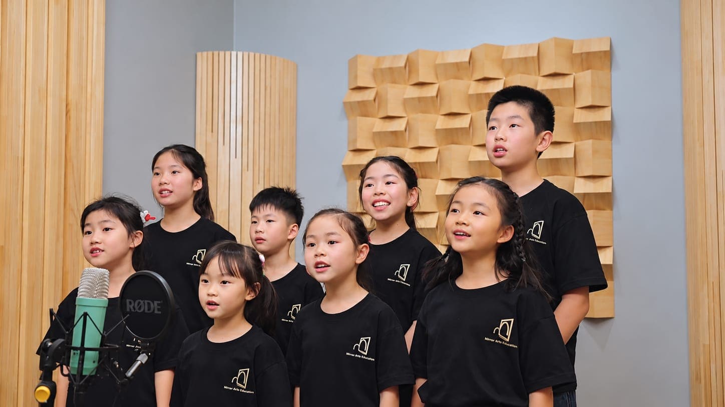Eight children in Mirror T-shirts singing together in the recording studio, a large microphone in the foreground and acoustic panelling behind them