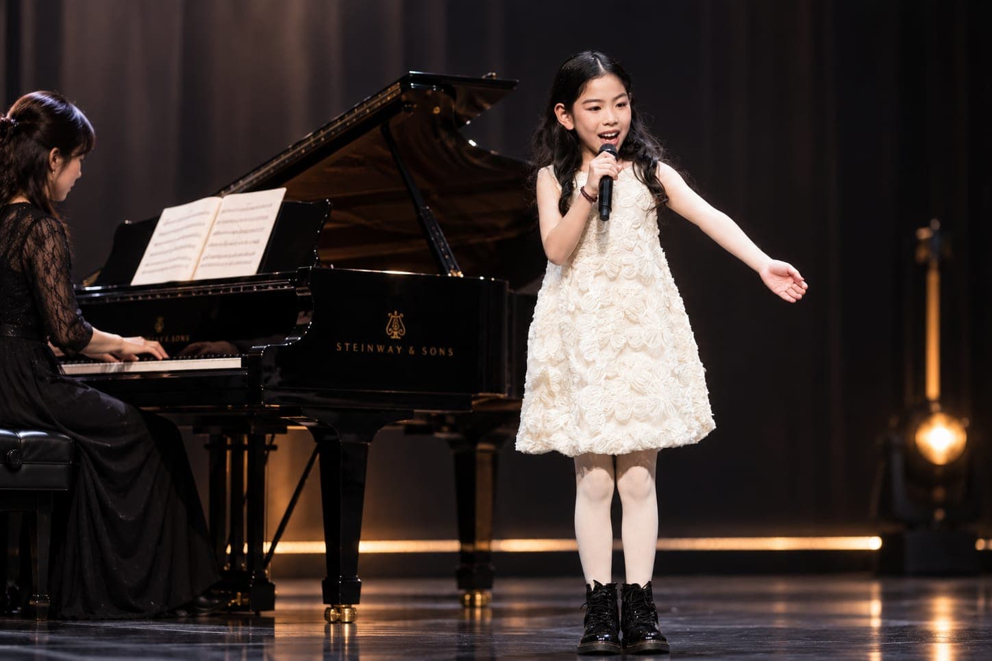 A girl singing at the microphone on a concert stage, her accompanist at a grand piano behind her