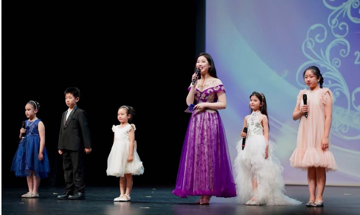 Five children beside their host on the gala stage: the girls in blue, white tulle and pink hold microphones, the boy in a black suit and the girl in a white dress do not